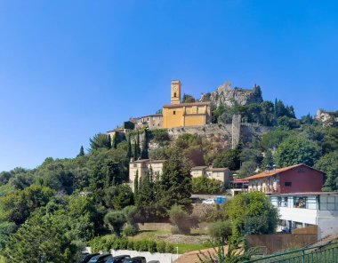 Eze, Fransa - 9 Temmuz 2024: Mavi gökyüzü, oteller ve tepenin aşağısındaki evler altındaki EZA kalesi ve kulesi ile kiliseye bakmak. Bir sürü yeşil ağaç.