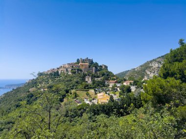 Eze, Fransa - 9 Temmuz 2024: Mavi gökyüzünün altındaki yeşil ormanlık tepenin üzerindeki tarihi köy