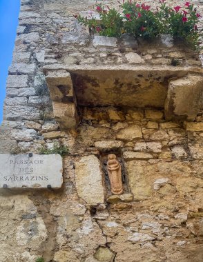 Eze, Fransa - 9 Temmuz 2024: Tarihi duvarda eski aşınmış Madonna heykeli. Sarrazins geçidi, patika, üzerinde kırmızı çiçekler var.