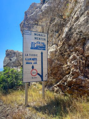 Eze, Fransa - 9 Temmuz 2024: Monako, Menton, La Turbie, Cenova ve Nice 'i gösteren yol işareti. Rocky Cliff arka planda. Mavi gökyüzü