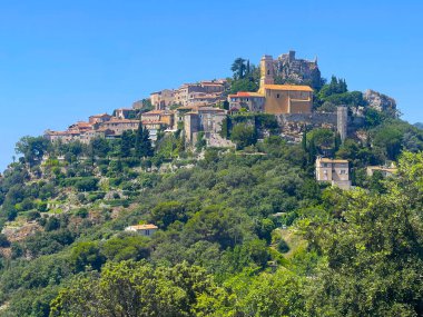 Eze, Fransa - 9 Temmuz 2024: Mavi gökyüzü, oteller ve tepenin aşağısındaki evler altındaki EZA kalesi ve kulesi ile kiliseye bakmak. Bir sürü yeşil ağaç.