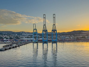 Marsilya, Fransa - 10 Temmuz 2024: Konteyner terminali ve limandaki vinçleri üzerinde gün doğumu. Şehir manzarası arkada. Uzakta dağ yamacına yazılmış uzun karakterlerin port adı