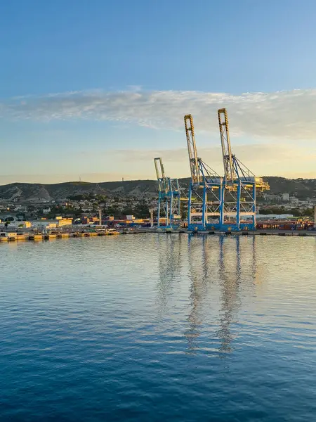 Marsilya, Fransa - 10 Temmuz 2024: Limandaki konteyner terminali ve vinçleri. Gündoğumu gökyüzü ve sağ taraftaki tüm öğeler güneş ışığıyla sarıya boyanmış.