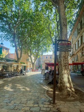 Aix-en-Provence, Fransa - 10 Temmuz 2024: Place des Martyrs de la Resistance. Başpiskoposun tarihi sarayı, şimdi müze, sonunda. Yeşil yapraklı ağaçlar barların dışında oturan insanları gölgeliyor.