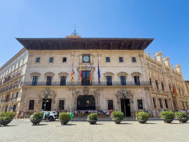 Palma de Mallorca, İspanya - 12 Temmuz 2024: Ayuntamiento, Placa de Cort AKA Palma Meydanı 'ndaki Şehir Konseyi tarihi binasında bayraklar ve saat bulunmaktadır. İnsanlar ve arabalar önde.
