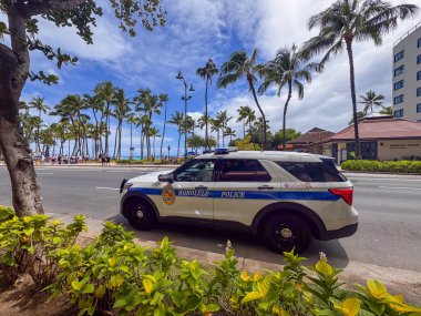 Honolulu, Oahu, Hawaii - July 9, 2025: Police car at Waikiki Beach with pedestrians on boardwalk under blue cloudscape. Palm trees and plants. Police base is across the street