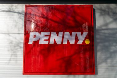 Parlak kırmızı tabela kalın beyaz harflerle Penny ismini içeriyor. Tabela, ağaç gölgeleriyle çevrili bir duvara asılmış, kentsel bir perakende atmosferi vurguluyor..