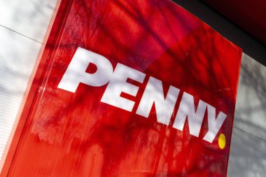Penny kelimesinin yer aldığı koyu kırmızı bir tabela, şehir mimarisinin bir zeminine dikkat çekiyor. Güneş ışığı gölgeler oluşturur, işaretin görünürlüğünü ve canlılığını arttırır..