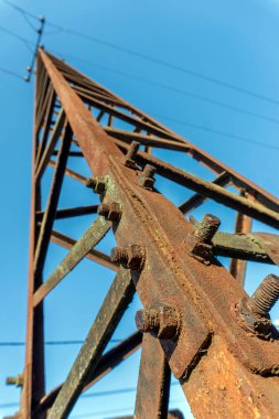 Bu güç hattı kulesi, belirgin bir pas ve yıpranma gösteriyor, açık mavi gökyüzüne karşı dimdik ayakta duruyor. Yapı, zamanın geçişini ve elementlere maruz kalışını vurgular.