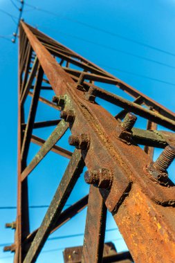 Parlak mavi gökyüzüne doğru yükselen paslı bir elektrik kulesinin detaylı bir görüntüsü. Yıpranmış metal ve belirgin civatalar, öğleyin güneş ışığı altında yapının yaşını ve çürümesini vurguluyor..