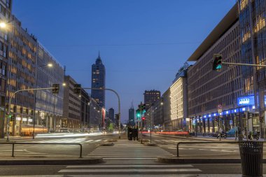 Milan, İtalya - 4 Nisan 2025: Aydınlatılmış binalar, arabalar ve yayalar alacakaranlıkta yol alırken hareketli bir cadde çiziyor. Atmosfer canlı şehir ışıklarıyla büyüleyici bir manzara yaratıyor. Milano şehrinde.