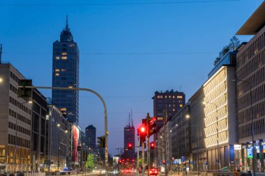 Milan, İtalya - 4 Nisan 2025: Aydınlatılmış binalar, arabalar ve yayalar alacakaranlıkta yol alırken hareketli bir cadde çiziyor. Atmosfer canlı şehir ışıklarıyla büyüleyici bir manzara yaratıyor. Milano şehrinde.