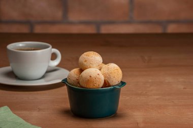 Cheese breads in a green ramekin with a cup of coffee on wooden table and bricks wall background.