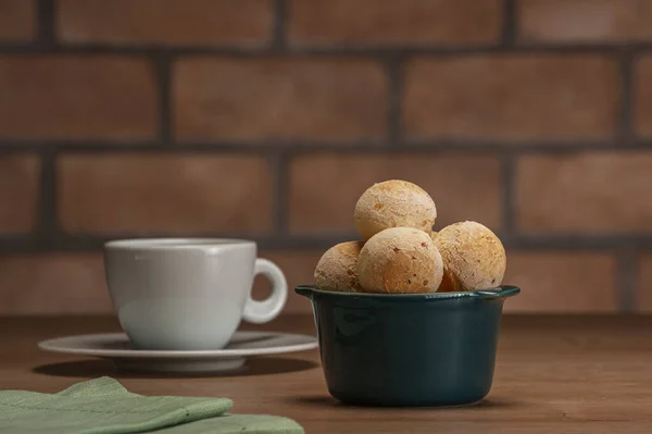 Cheese breads in a green ramekin with a cup of coffee on wooden table and bricks wall background.