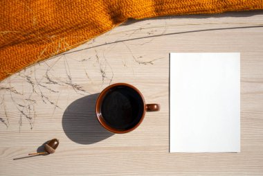 Brown cup of hot coffee, acorn, blank card empty copy space for mock up, orange knitted sweater on a light wooden surface. Flat lay, top view. Autumn season template. Authors Day. Coffee break