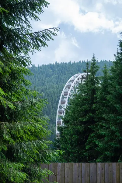 stock image Fragment of ferris wheel against the blue sky with clouds and green tall coniferous trees of the Carpathian forest. Summer ecological vacation