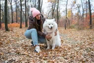 Sonbahar parkında Samoyed köpeğiyle gülümseyen güzel kız. Esmer kadınlar pofuduk güzel köpekle eğleniyor. Kız köpeğe sarılıyor. Bir köpeği öp.
