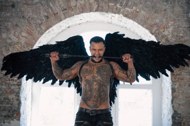 Kanatlı yakışıklı adam. Siyah kanatlı erkek melek. Kırbaçlı, kaslı, üstsüz bir adam. Dövmeli, vahşi, yakışıklı bir adam. Kaslı atletik seksi erkek 