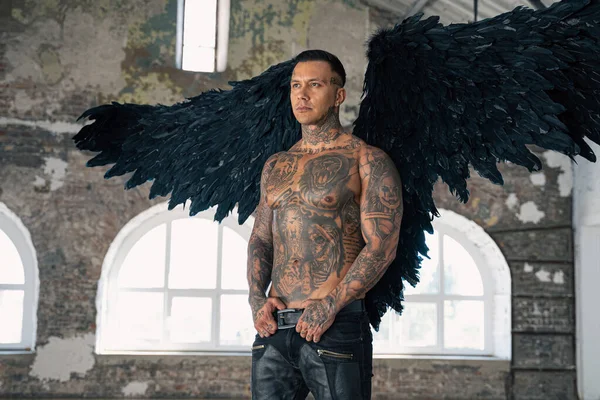 angel man with black wings, stand posing at camera. man fall from heaven, angel with muscular body gained freedom.  tattooed hot man. tattooed black angel. dark angel in leather