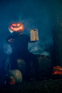 Headless man holds Jack O'Lantern Halloween pumpkin, hold devil's contract. Trick and treat. orange glowing carved face instead of head on countryside road in autumn foggy forest.