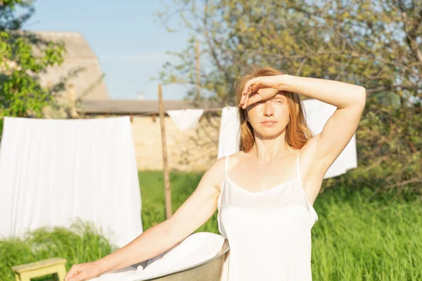 stock image beautiful red girl in nightie hanging laundry outdoors. village woman working in countryside.Cute girl in dress washing white clothes in metal basin in backyard, hanging laundry on clothesline