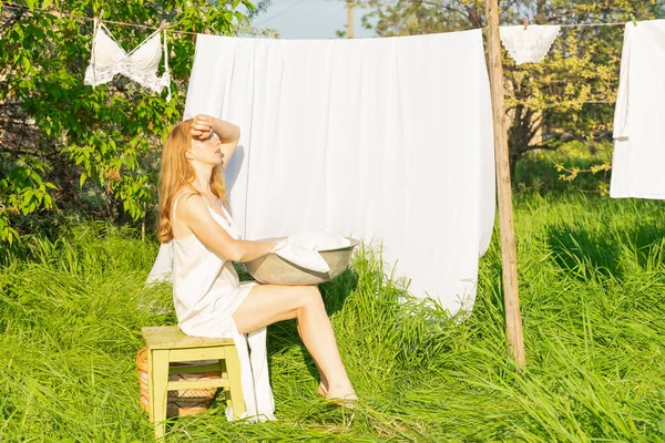 stock image beautiful red girl in nightie hanging laundry outdoors. village woman working in countryside.Cute girl in dress washing white clothes in metal basin in backyard, hanging laundry on clothesline