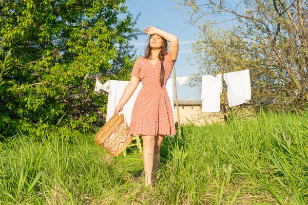 stock image Young woman hanging laundry outdoors. Beautiful girl in red polkadot dress. working in countryside. Cute brunette girl in dress washing white clothes in metal basin in backyard. village girl