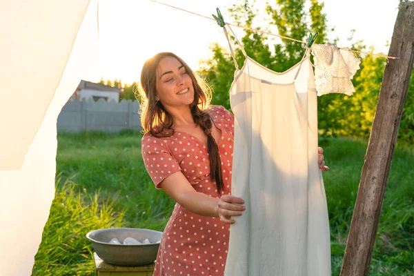 stock image Young woman hanging laundry outdoors. Beautiful girl in red polkadot dress. working in countryside. Cute brunette girl in dress washing white clothes in metal basin in backyard. village girl