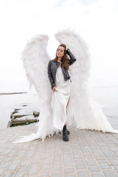白い翼を持つ美しい若い女性 革のジャケットと白い絹のドレスで白い天使 聖なる女 — ストック写真