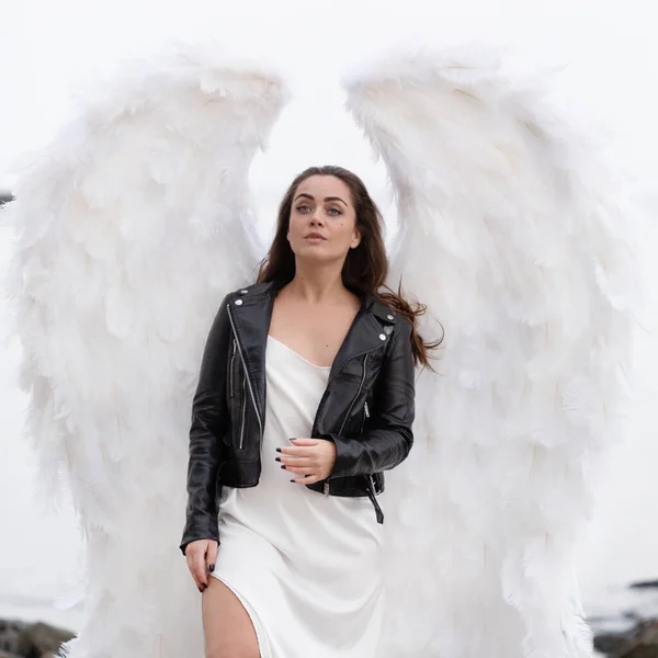 白い翼を持つ美しい若い女性 革のジャケットと白い絹のドレスで白い天使 聖なる女 — ストック写真