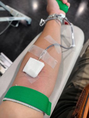 Kan bağışı yapan birinin yakın plan görüntüsü. İğne koluna sokuluyor, tıbbi bantla korunuyor ve kan toplama tüpüne bağlanıyor. Yeşil turnike uygulanır. Görüntü sağlık, yardım, tıbbi prosedürleri temsil ediyor.