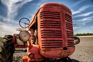 Mavi gökyüzünün altında dağınık bulutlu eski, kırmızı bir traktörün yakın görüntüsü. Traktörde aşınma ve pas izleri var. Arkaplanda kırsal ya da tarımsal bir ortam izlenimi veren açık bir alan var.