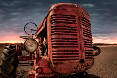 Eski, kırmızı bir traktör, görünür pas ve yıpranma izleri var. Traktör, gün doğumu ya da gün batımı gibi görünen bir alanda konumlandırılmış. Teknoloji ve doğa arasındaki zıtlık, nostalji ve zamanın geçişini çağrıştırıyor..