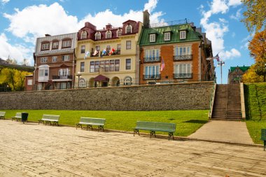 Eski Quebec 'ten, mimari stil ve renklerin karışımından oluşan büyüleyici bir binaya odaklanmış bir sahne. Ön planda yeşil banklar var ve bina birden fazla bayrakla süslenmiş..