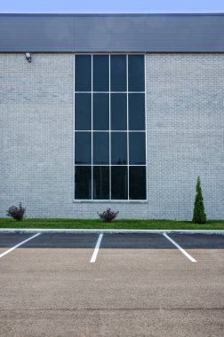 Merkezinde büyük bir pencere olan modern bir binanın dış cephesi. Ayrıca ön planda açıkça işaretlenmiş park yerleri olan bir park yeri de var. Ticari veya ofis binası, mimari tasarımı ve peyzajını vurguluyor.