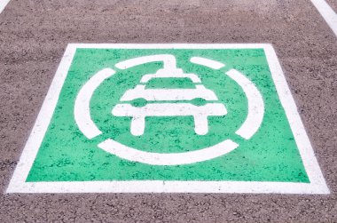 Sembol fişi olan bir arabayı gösteriyor, bu da elektrikli araçlar için ayrılmış bir park yeri olduğunu gösteriyor. Asfaltlı bir yüzeye beyaz çizgili ve içinde beyaz bir sembol bulunan yeşil bir kare..