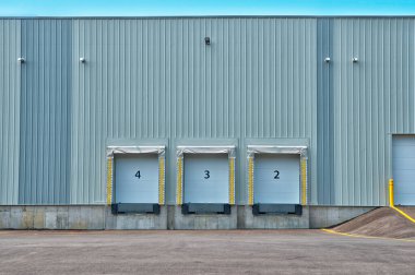 Üç endüstriyel yükleme rıhtımı olan bir binanın yan görüntüsü. Her rıhtımın kapalı bir mavi rulo kapısı var ve tabanında uzatılabilir bir rıhtım düzleştiricisi var..