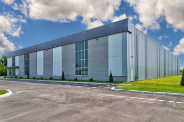 Metal kaplamalı modern endüstriyel bina, büyük cam pencereler ve parlak mavi gökyüzüne karşı kurulmuş geniş bir otopark. Ticari gayrimenkul, iş ve sanayi temaları için ideal