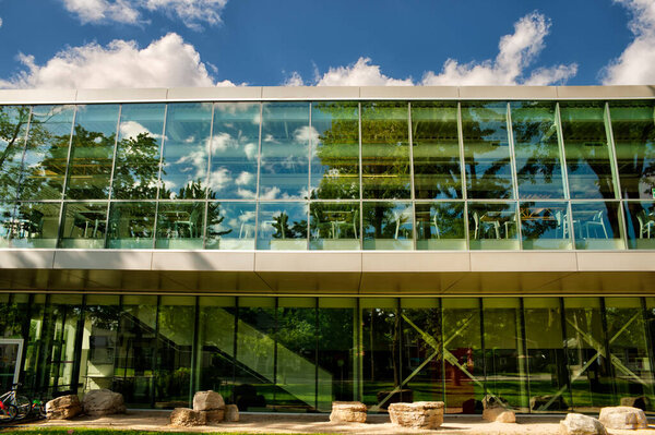 Современное стеклянное здание, отражающее деревья и голубое небо, демонстрирует сочетание природы и современной архитектуры. Дизайн подчеркивает прозрачность, свет и гармонию с окружающей средой.