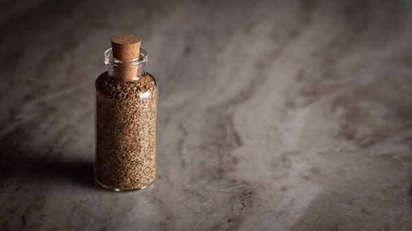 A small glass bottle filled with organic Carom seeds (Trachyspermum ammi) or Ajwain is placed on a marble background.