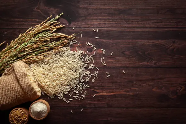 Stock image Organic whole Rice and Rice Bran (Oryza sativa) spilled out from a laying jute bag, isolated on a dark wooden background. Top view.