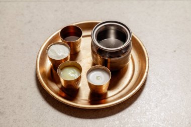 A copper plate with panchamrit items in brass glasses (milk, honey, ghee, curd) alongside water in a steel lota, prepared for the Krishna Janmashtami celebration. clipart