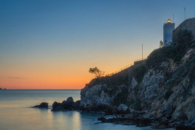 Talamone kayalık plajı ve deniz feneri gün batımında. Maremma, Grosseto ili, Toskana bölgesi, İtalya.