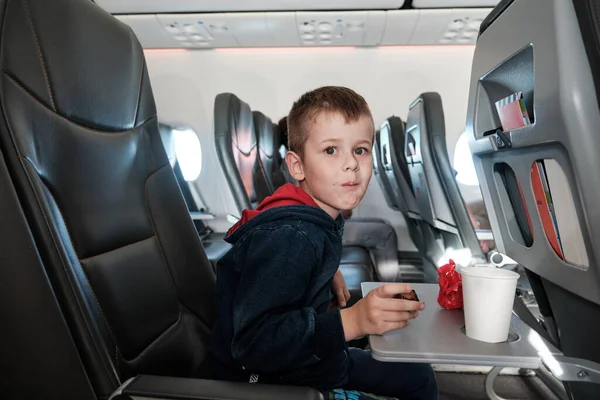 A little boy on board the plane eats chocolate candies while drinking water. Six years old boy traveling by an airplane.