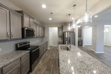 Paslanmaz çelik aletleri, granit tezgahları ve şık dolapları olan modern bir mutfak. Geniş bir ada ve cam kolye ışıklarıyla açık alan. Yatırım, kiralama veya ev sahipliği kavramı.