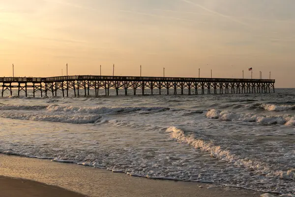Oak Island İskelesi, Kuzey Carolina 'da parlayan gündoğumu gökyüzünün altında Atlantik Okyanusu' na doğru uzanırken dalgalar kıyı boyunca çarpar..