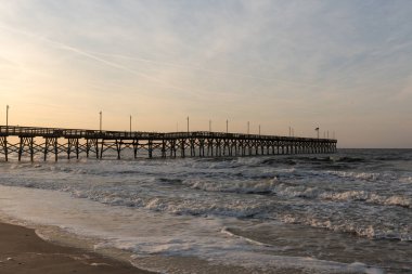 Oak Island İskelesi, Kuzey Carolina 'da parlayan gündoğumu gökyüzünün altında Atlantik Okyanusu' na doğru uzanırken dalgalar kıyı boyunca çarpar..