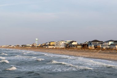 OAK ISLAND, NC, ABD - 13 Mart 2025: Okyanus dalgaları yumuşak mavi gökyüzü altında renkli evlerle dolu kumlu kıyılara doğru yuvarlanıyor.