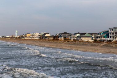 OAK ISLAND, NC, ABD - 13 Mart 2025: Okyanus dalgaları yumuşak mavi gökyüzü altında renkli evlerle dolu kumlu kıyılara doğru yuvarlanıyor.