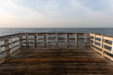 Yıpranmış ahşap bir balıkçı iskelesi okyanusun üzerine uzanır sakin bir gökyüzünün altında, parmaklıklar boyunca görünen balıkçılar için numaralandırılmış istasyonlar vardır..